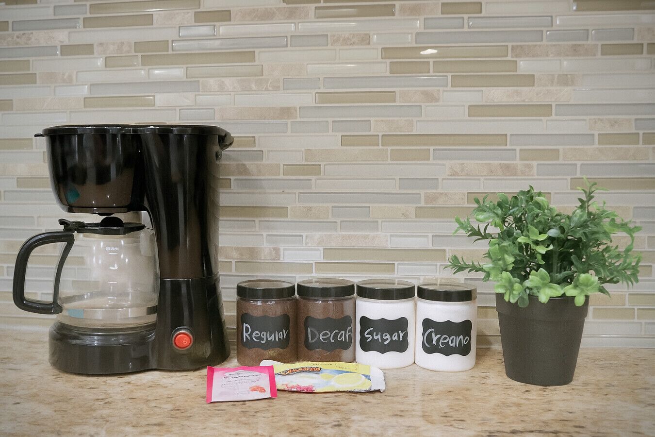 A kitchen counter with a coffee maker, four labeled jars, a potted plant, and a packet of sugar.