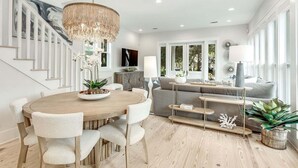 A modern living room with a round wooden dining table, white upholstered chairs, a grey sofa, and a staircase with white railings. - Coastal Corner Cottage (Santa Rosa Beach)