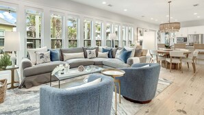 A modern living room with a sectional sofa, a glass coffee table, and a dining area with a chandelier. - Coastal Corner Cottage (Santa Rosa Beach)