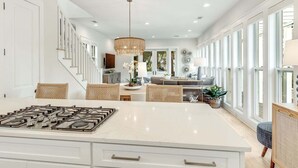 A spacious kitchen with a large island, a dining area with wicker furniture, and a view of the outdoors through large windows. - Coastal Corner Cottage (Santa Rosa Beach)
