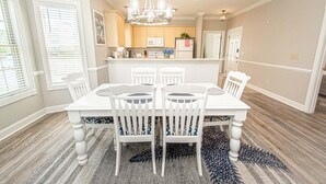 Un coin repas avec une table et des chaises blanches, un lustre et une cuisine en arrière-plan.