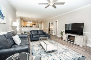 A modern living room with a leather sofa, a glass coffee table, a flat-screen TV, and a kitchen in the background. - Magnolia Pointe Luster Leaf 4891-205 (Myrtle Beach)
