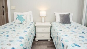 Une chambre avec deux lits, chacun agrémenté d’un oreiller blanc et d’un oreiller bleu orné d’un motif représentant une tortue de mer. Une table de chevet blanche avec une lampe se trouve entre les deux lits.