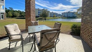 Une terrasse extérieure avec une table et des chaises, un pilier en briques, et une vue sur un lac et des bâtiments en arrière-plan.