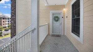 A covered porch with a white railing, a door with a wreath, and a window with blinds. - Magnolia Pointe Luster Leaf 4890-301 (Myrtle Beach)