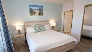 A bedroom with a wooden bed, two bedside tables with lamps, a beach-themed pillow, and a door to another room. - Magnolia North 4839-203 (Myrtle Beach)