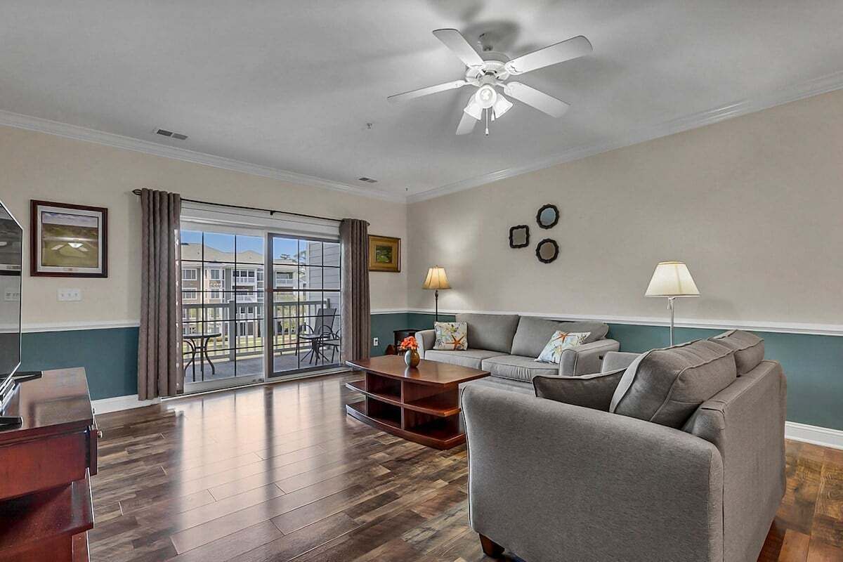 A living room with a grey sofa, a wooden coffee table, and a ceiling fan. There is a window with curtains, a framed picture on the wall, and a lamp on a side table.