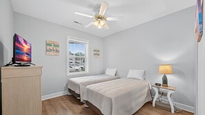 Une chambre avec deux lits, un ventilateur de plafond, un bureau avec un ordinateur, une fenêtre donnant sur l’extérieur et une table de chevet avec une lampe.