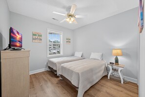 A bedroom with two beds, a ceiling fan, a desk with a computer, a window with a view of the outside, and a nightstand with a lamp. - Magnolia Place 4789-104 (Myrtle Beach)