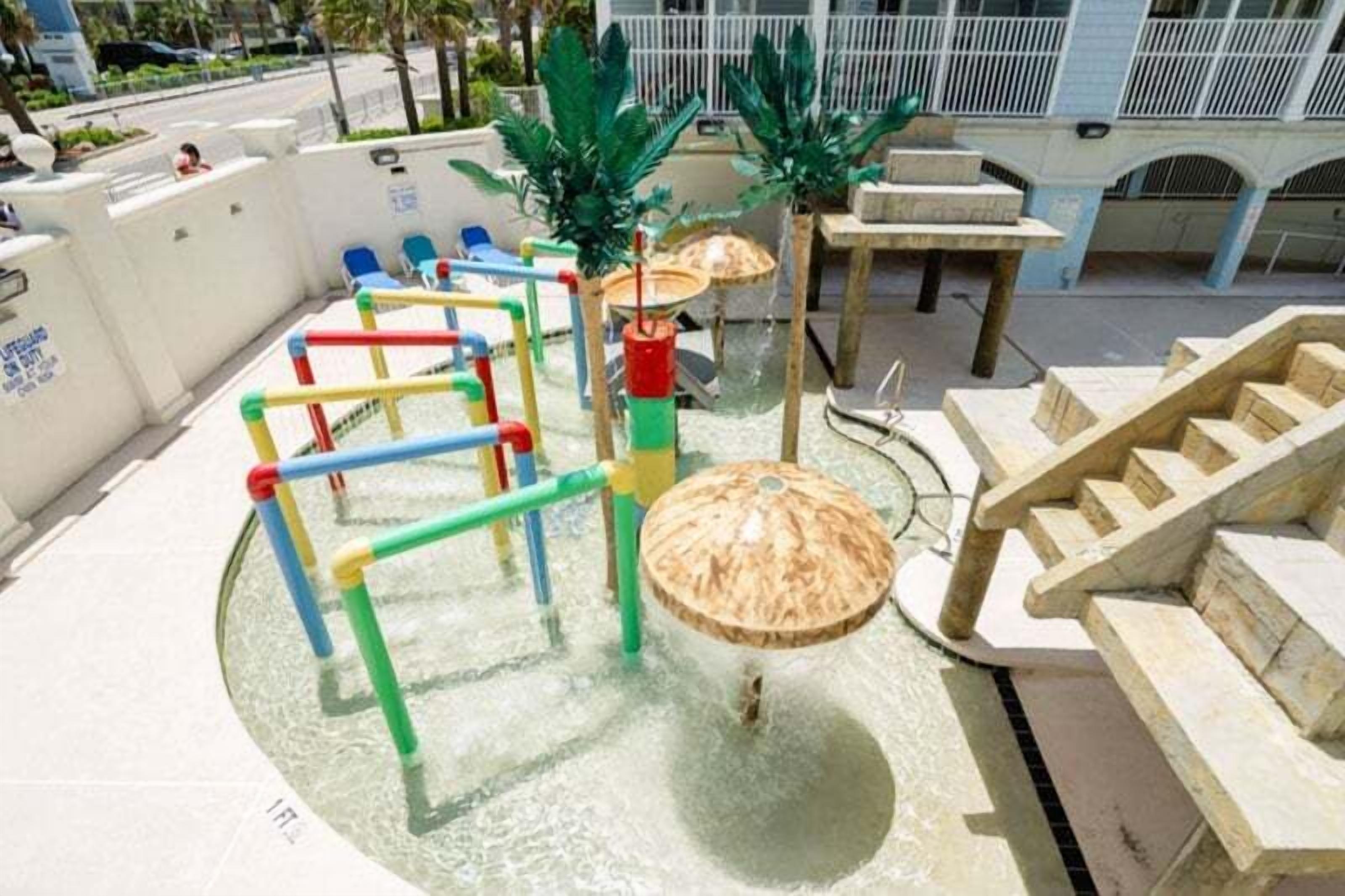 Un espace de jeux pour enfants avec une table à eau, des structures colorées et un escalier en bois.