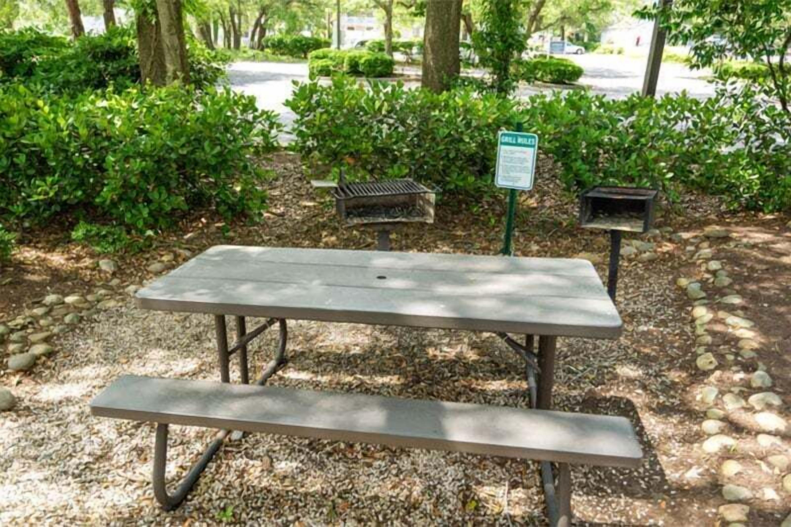 A picnic table in a park with a grill and a trash bin nearby.