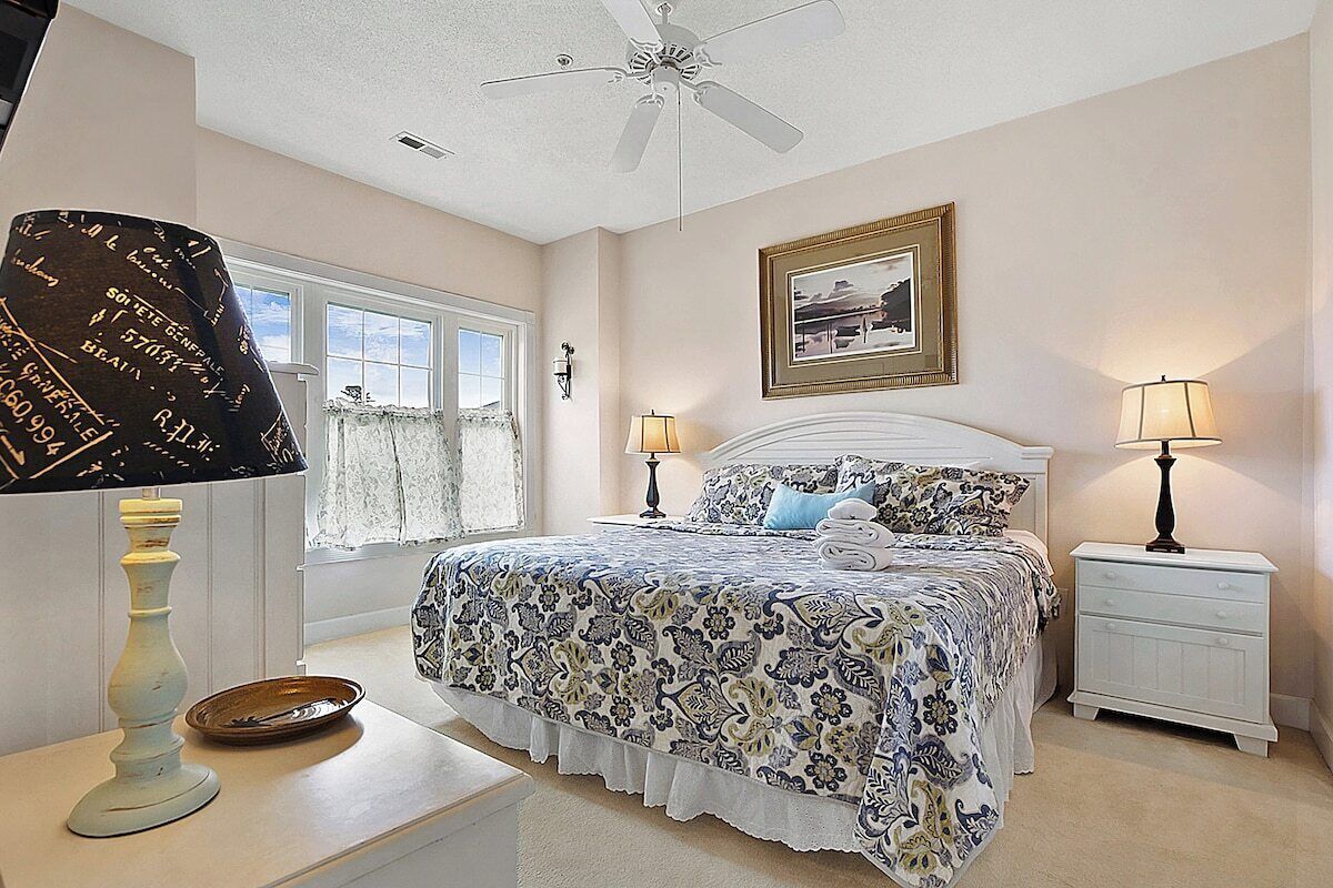 A bedroom with a bed, two bedside tables, a ceiling fan, a window, and a framed picture on the wall.