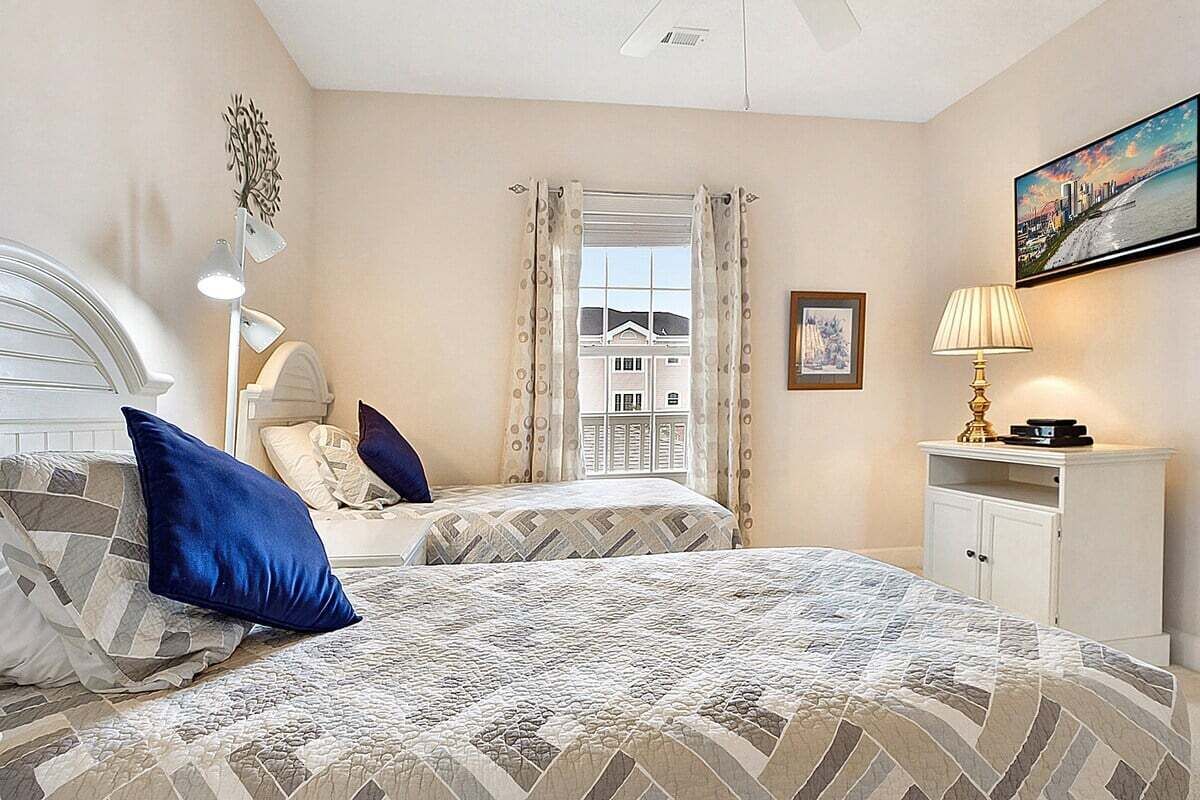 A bedroom with two beds, a window with curtains, a painting on the wall, and a lamp on a nightstand.