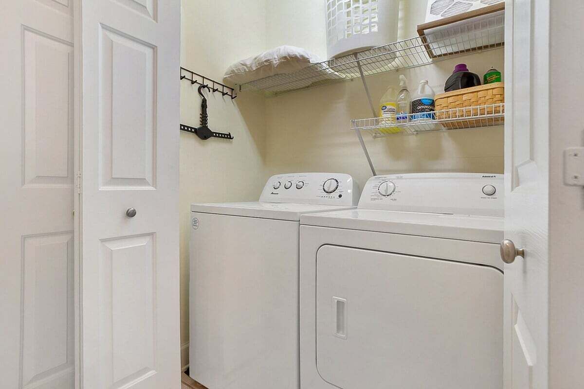 A laundry area with a front-loading washer and dryer, a drying rack, and shelves with laundry supplies.