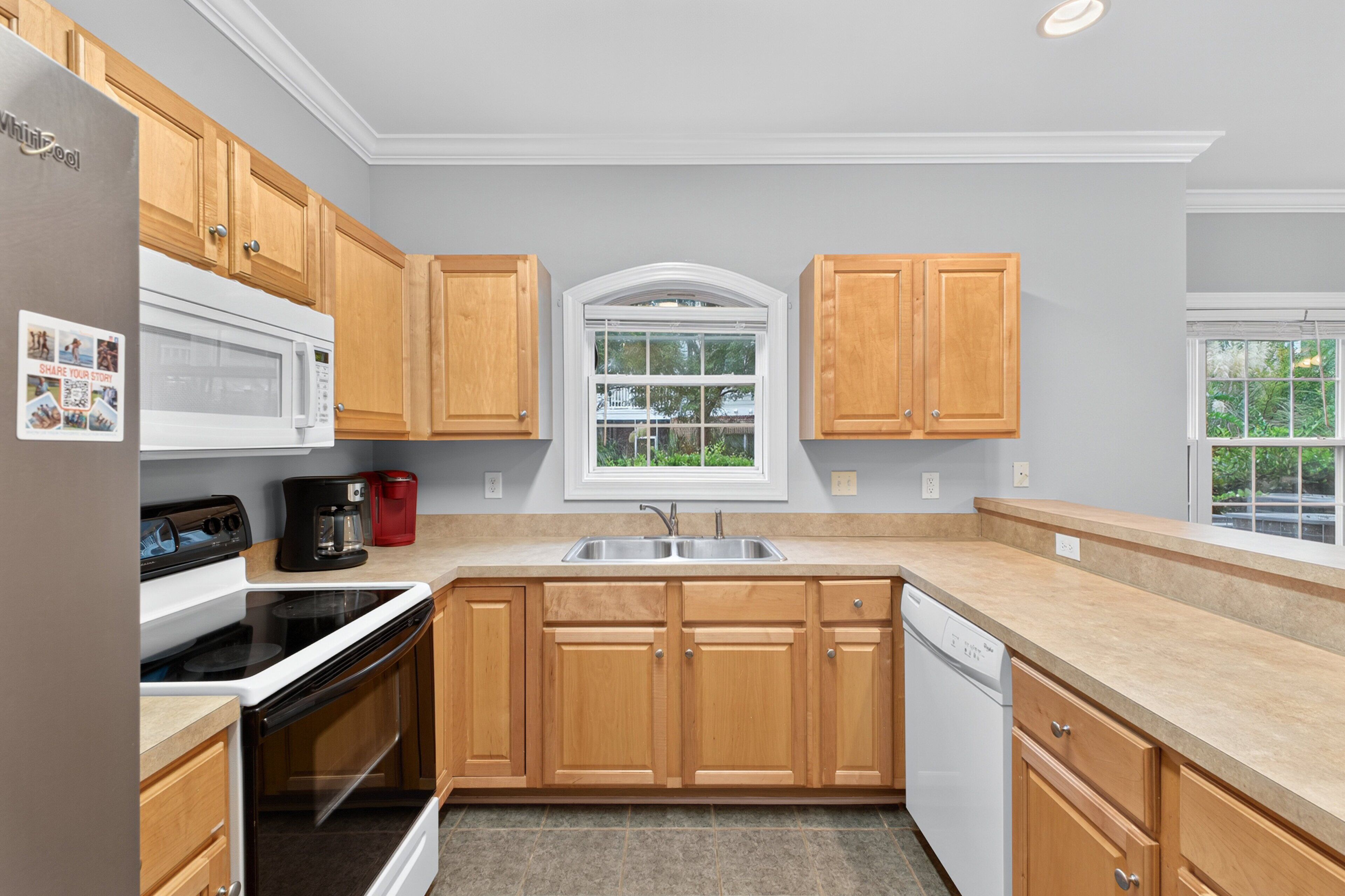 A kitchen with wooden cabinets, a white refrigerator, a microwave, a stove, a dishwasher, and a window with a view of greenery.