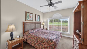 A bedroom with a bed, nightstand, lamp, ceiling fan, window, and a view of a green landscape. - Magnolia Place 4701-104 (Myrtle Beach)