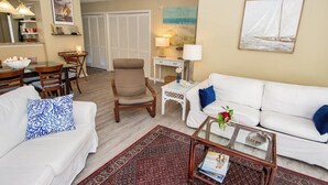 A living room with a white sofa, a coffee table, and a dining area with a table and chairs. - West Hyde Park 2E (Myrtle Beach)