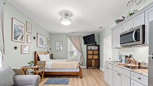 Ein Schlafzimmer mit Bett, Nachttisch und Fernseher. Das Zimmer verfügt über ein Fenster mit Vorhängen, einen Deckenventilator und eine Küchenzeile mit Mikrowelle und Spüle.