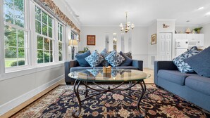 Ein Wohnzimmer mit einem Glastisch, blauen Sofas, gemusterten Kissen und einem großen Fenster mit Blick auf Grünflächen.