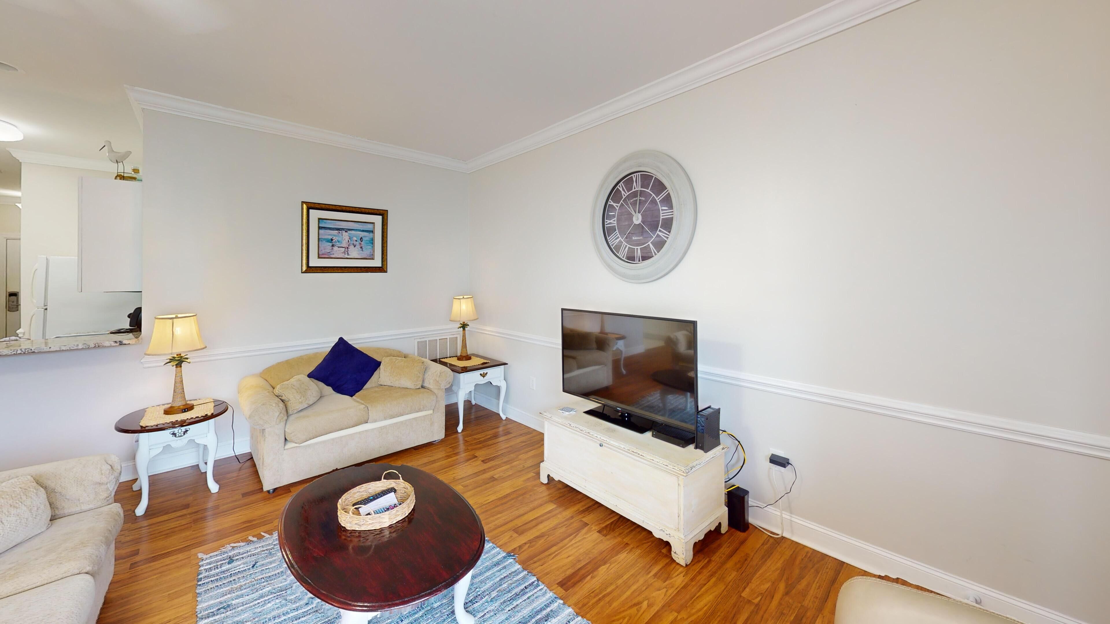 A living room with a beige sofa, a wooden coffee table, a TV, and a wall clock.