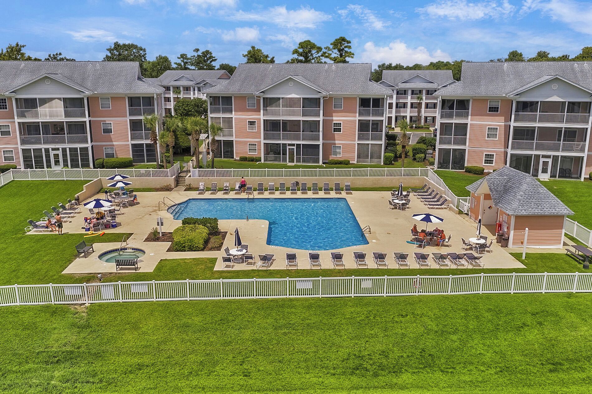 A hotel with a central swimming pool surrounded by lounge chairs and umbrellas, a fenced patio area, and a small building with a sloped roof.