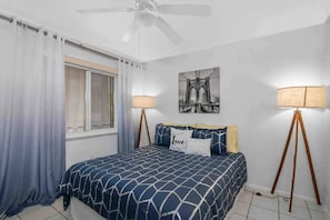 Ein Schlafzimmer mit Bett, Deckenventilator, Fenster mit Vorhängen, Wandlampe und einem gerahmten Bild.