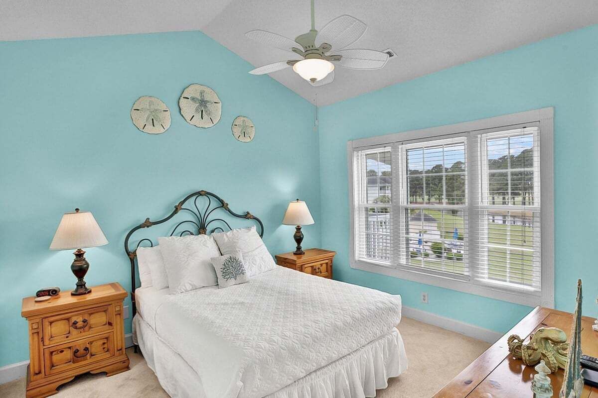 A bedroom with a bed, two nightstands, a ceiling fan, a window with blinds, and three wall clocks.
