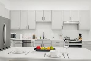 A modern kitchen with white cabinets, a stainless steel refrigerator, a microwave, a stove, and a dining area with a white table set for two. - Relaxing Modern Stay - Workerfriendly w/ Pool+gym (Sherman)
