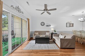 Ein geräumiges Wohnzimmer mit einer großen Schiebeglasdoor zu einer Terrasse, einem Deckenventilator, einer Uhr und einem Essbereich mit Tisch und Stühlen.