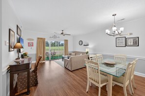 A dining area with a glass table, wooden chairs, a ceiling fan, and a view through a glass door to an outdoor space. - Magnolia Place 4701-103 (Myrtle Beach)