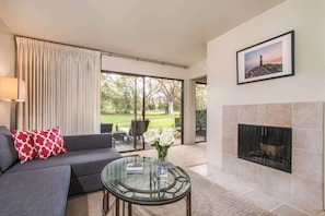 Un salon moderne avec une table basse en verre, un canapé orné de coussins rouges et blancs, une cheminée et une grande fenêtre donnant sur un jardin.
