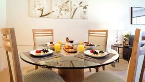 Un comedor con una mesa de vidrio rodeada de sillas, que ofrece productos para el desayuno como pasteles, frutas y zumo.