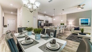 A modern kitchen with stainless steel appliances, a dining area with a white table set for four, and a living room with a sofa and a painting on the wall. - Peq3141 (Kissimmee)