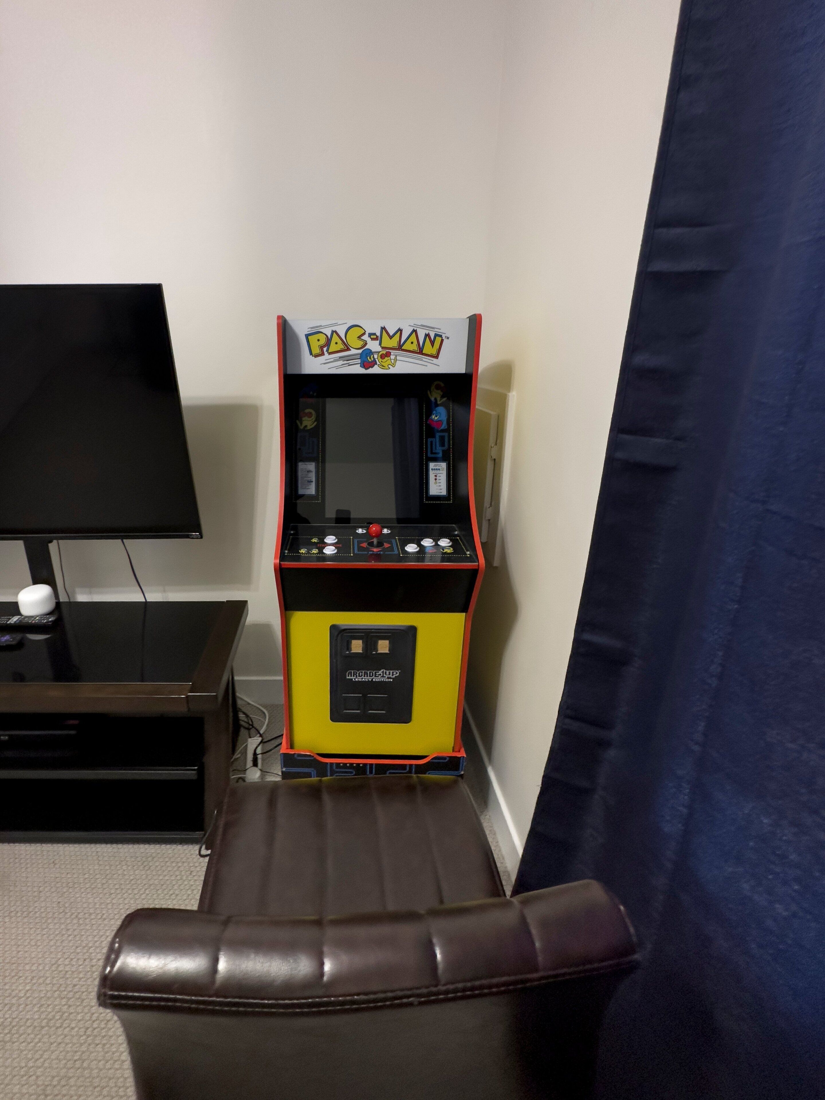 Ein Pac-Man-Automaten im Raum mit einem Fernseher und einem Ledersessel.