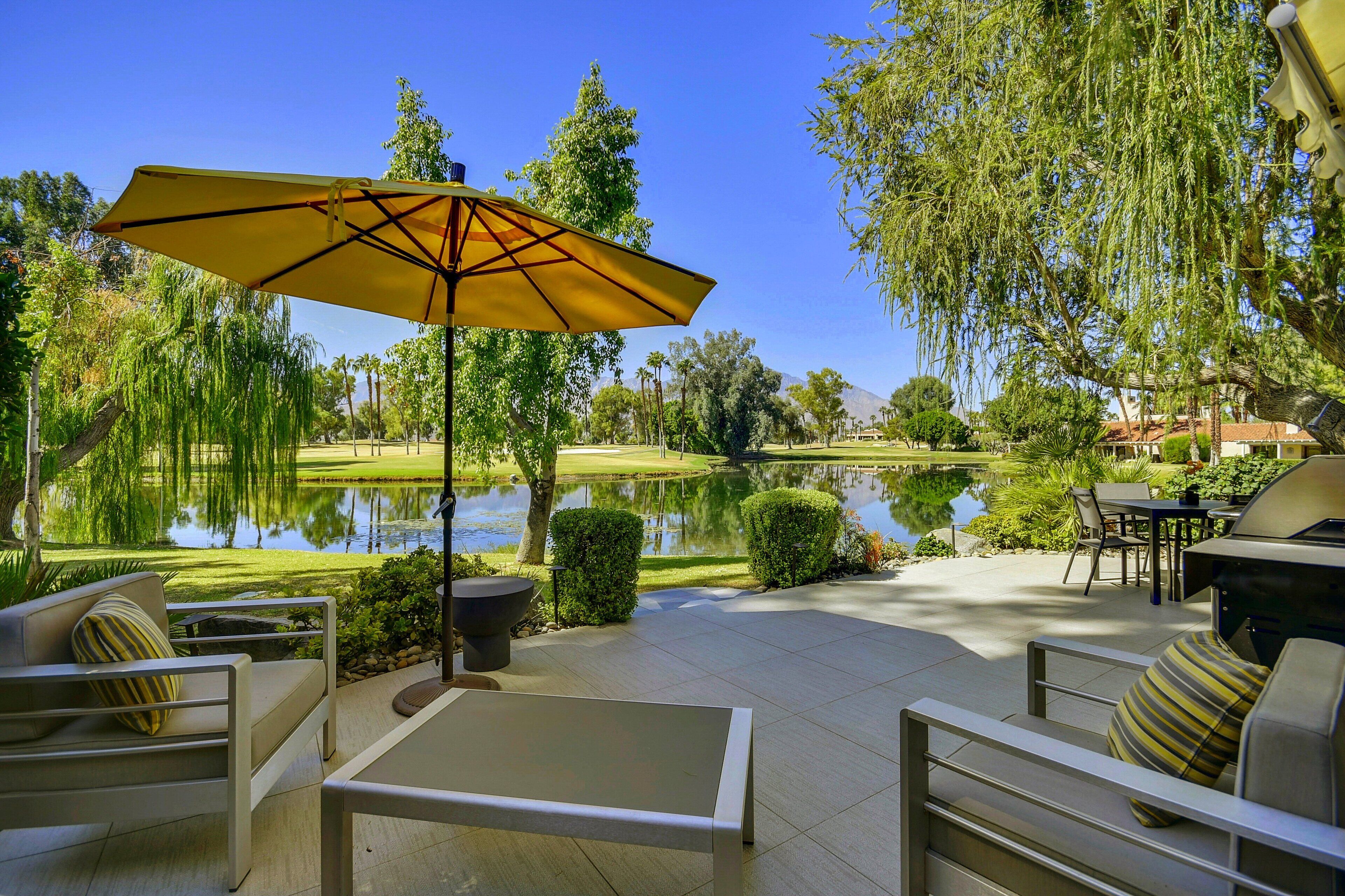 An outdoor patio with a table and chairs, a yellow umbrella, and a view of a lake and trees.