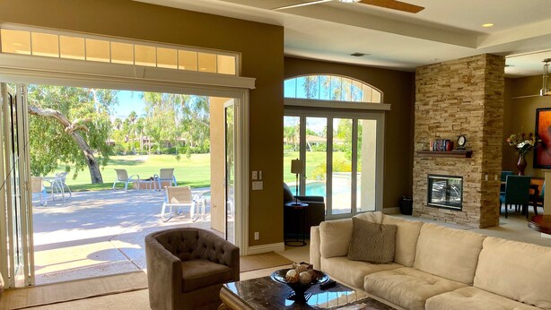 A spacious living area with a large sliding glass door leading to a patio, a fireplace, and a view of a golf course. - Villa Bella (Rancho Mirage)