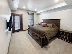 A bedroom with a large bed, a nightstand, a dresser, a TV mounted on the wall, and two windows with blinds.