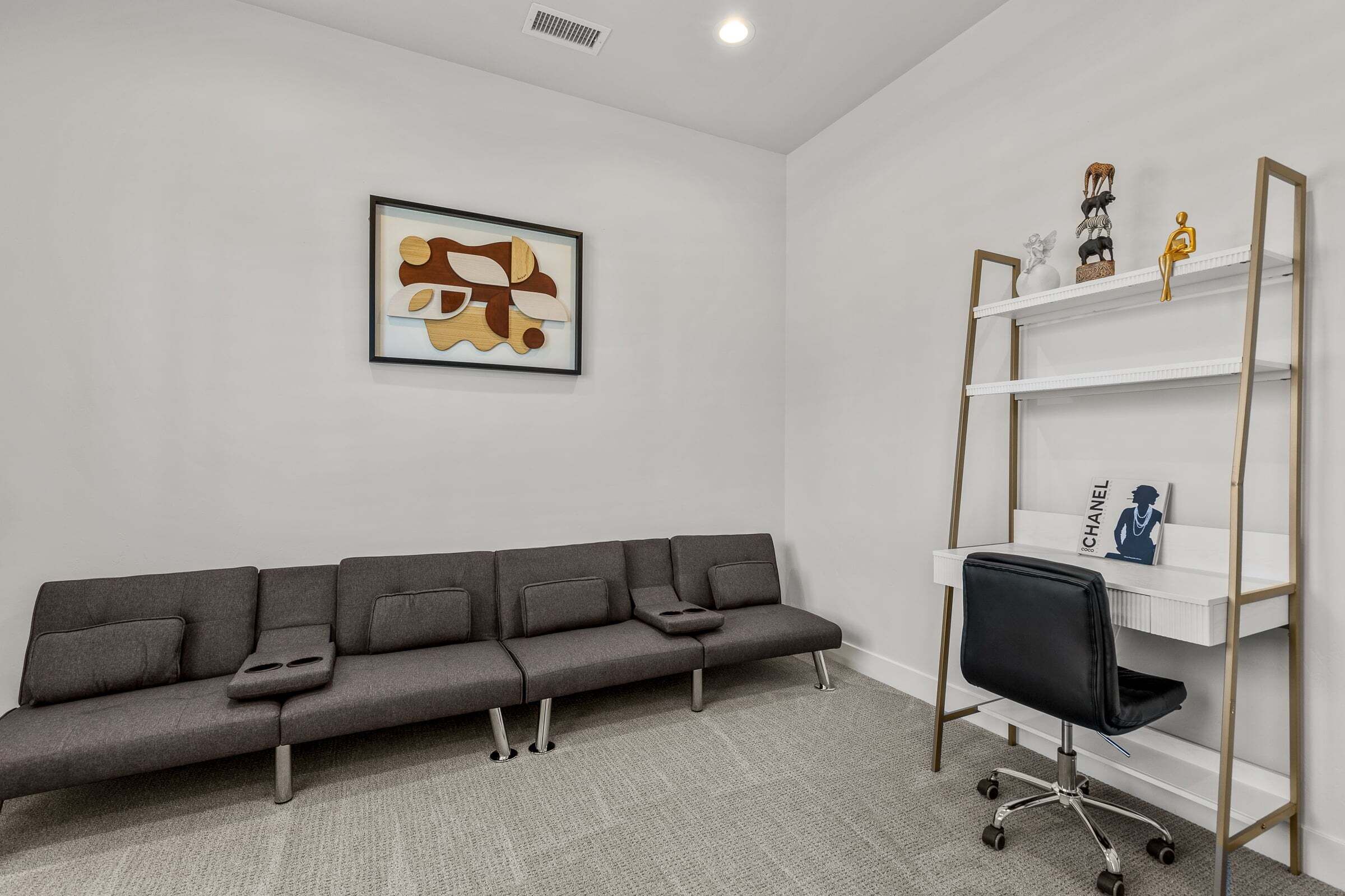 Une pièce moderne avec un canapé gris, un fauteuil de bureau noir, une étagère blanche agrémentée d’objets décoratifs et une œuvre d’art abstraite encadrée accrochée au mur.