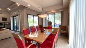 A dining area with a wooden table and red chairs, a chandelier, and large windows with a view of greenery. - Double Eagle (Rancho Mirage)