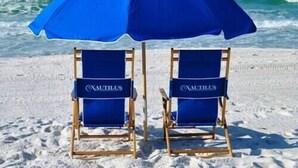 Zwei Strandstühle mit 'NAUTILUS'-Branding an einem sandigen Strand, im Hintergrund das Meer und Wellen.