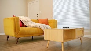A yellow sofa with a white throw blanket and orange pillows, a wooden coffee table, and a window with blinds. - Kingbed, Resort Style Pool, Gym, Neartheheights (Houston)