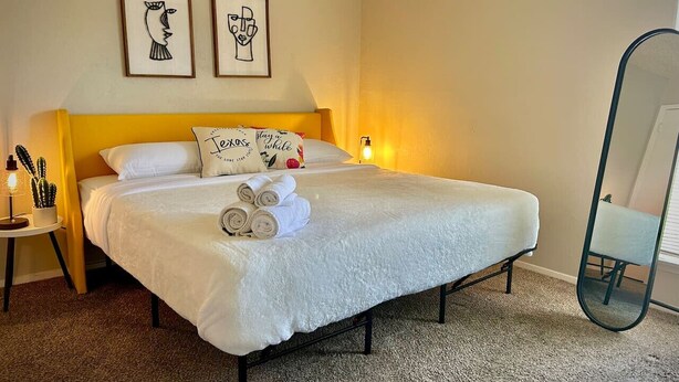 Een net opgemaakt bed met opgerolde handdoeken, een bedlampje en twee ingelijste kunstwerken aan de muur.