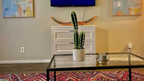 Een woonkamer met een flatscreen-televisie waarop 'NETFLIX' staat, een witte kast, een glazen salontafel met een cactus en abstracte schilderijen aan de muren.