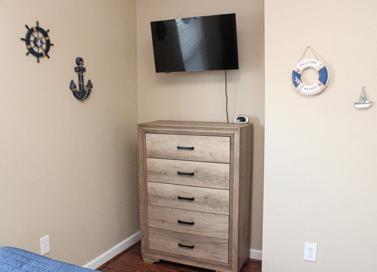 A bedroom with a wooden dresser, a television mounted on the wall, and nautical-themed wall decor.