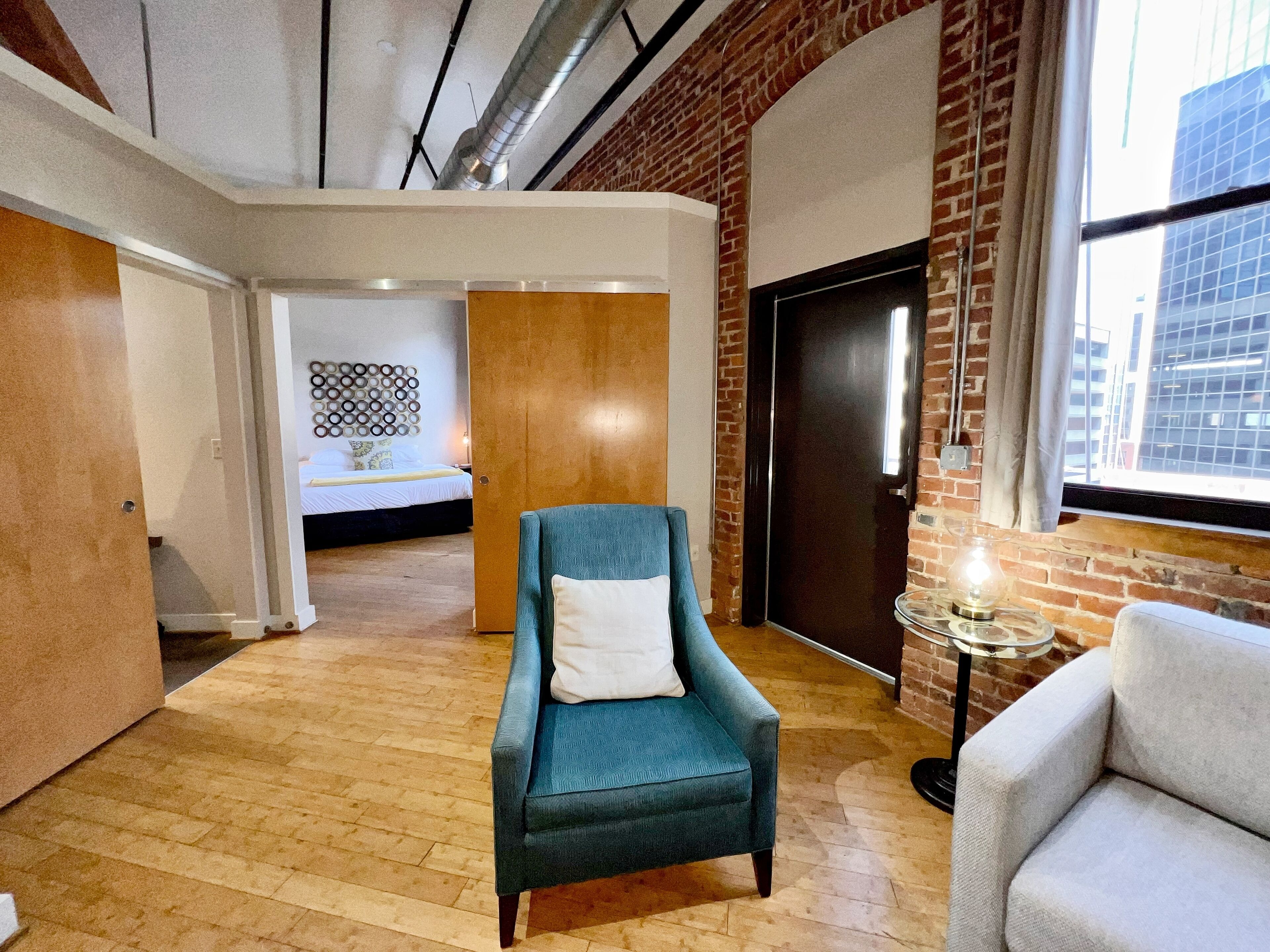 A modern hotel room with a wooden floor, a blue armchair with a white pillow, a white armchair, a small round table with a lamp, and a view of the city through a large window.
