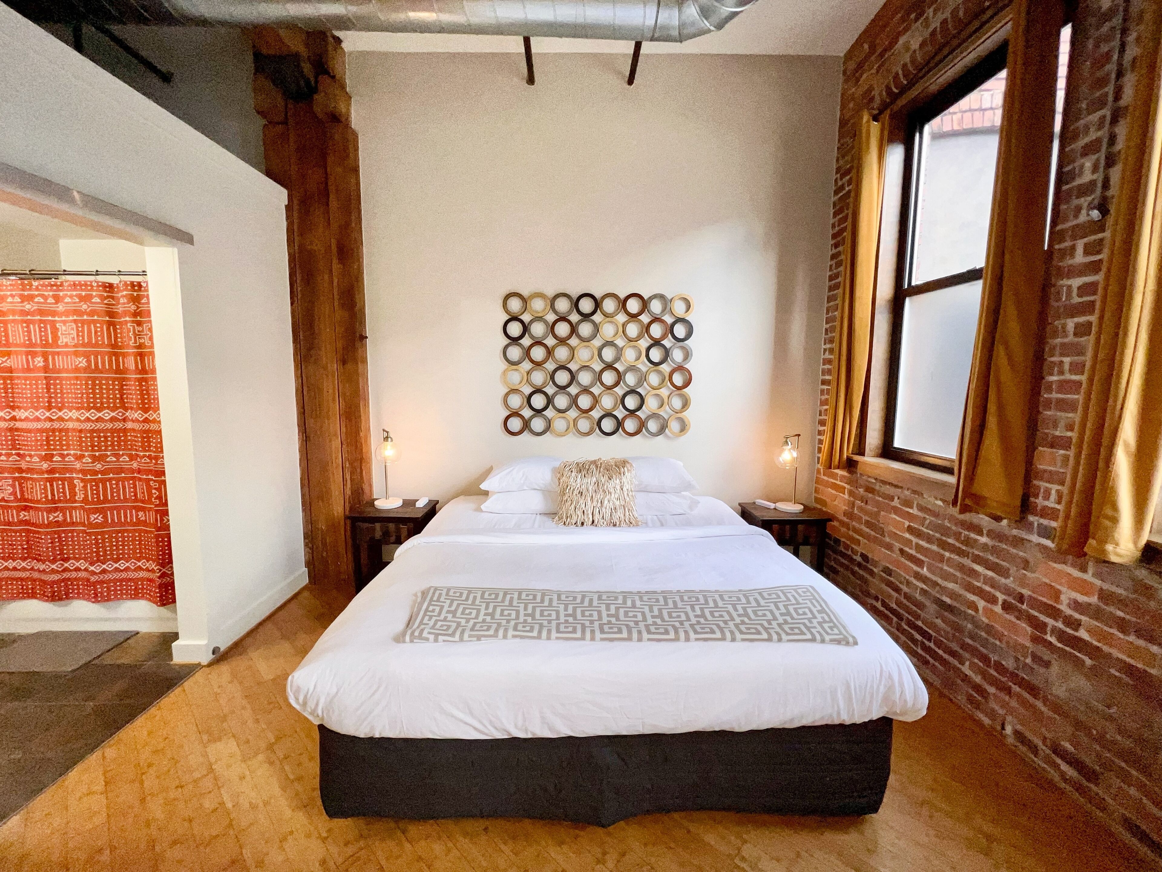 A bedroom with a large bed, two bedside tables with lamps, a wall-mounted circular pattern, and a brick wall.