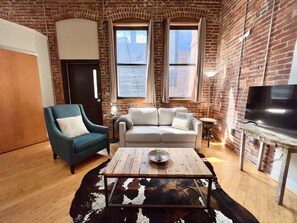 Un salon avec un mur en briques, deux fenêtres, un téléviseur, un fauteuil bleu, un canapé blanc et une table basse.