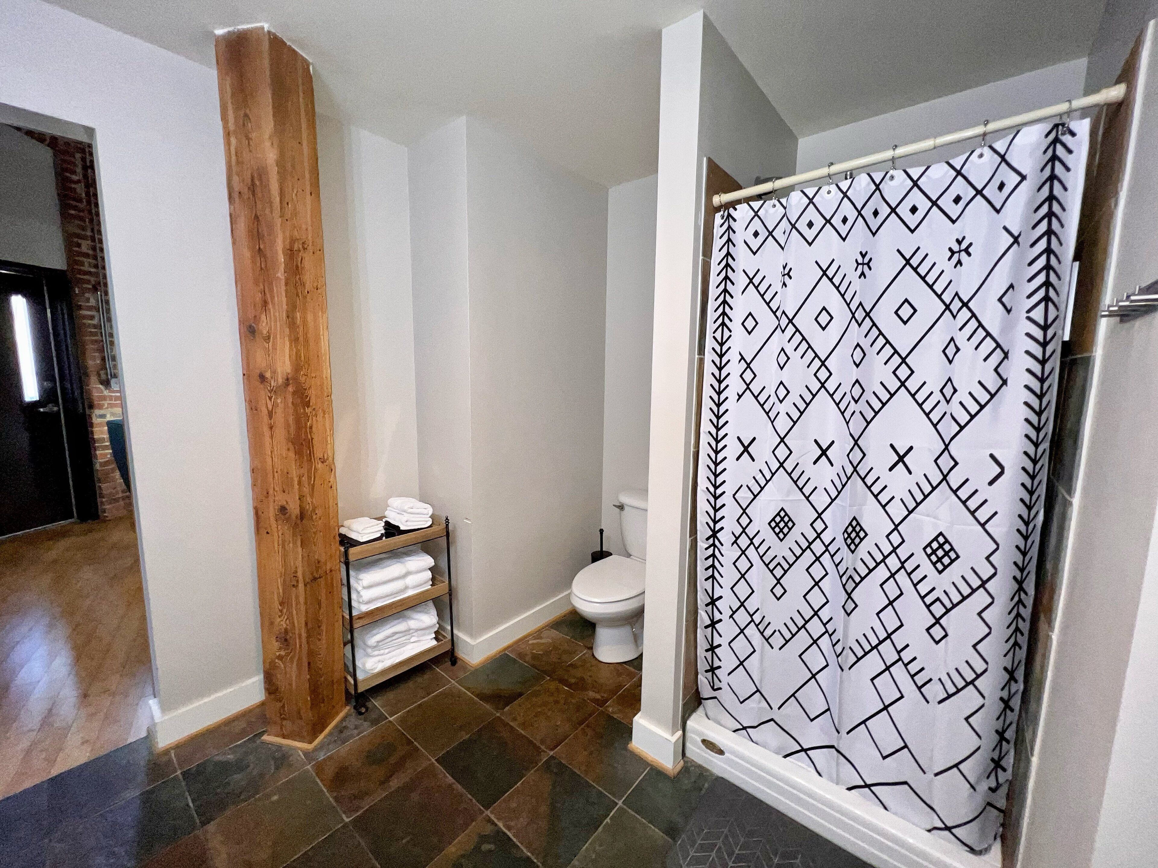 Une salle de bain avec un rideau de douche orné d’un motif géométrique, des toilettes et une étagère avec des serviettes pliées.