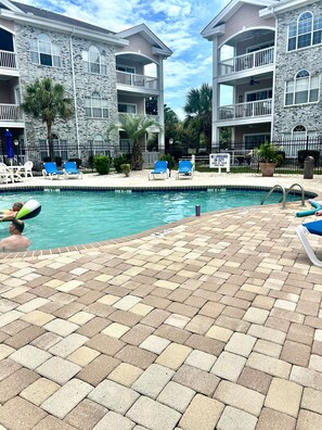 A pool area with lounge chairs, a sign stating 'NO LIFEGUARD ON DUTY', and a stone building with balconies. - Callaway Corner Luxe Pet-friendly 2BR Condo (Myrtle Beach)
