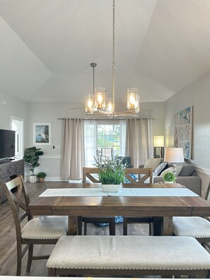 A dining area with a wooden table, cushioned chairs, a chandelier, and a view of the outdoors through large windows. - Callaway Corner Luxe Pet-friendly 2BR Condo (Myrtle Beach)